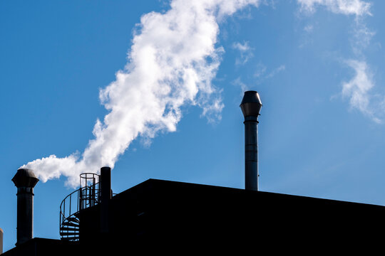Plume and steam from an industrial chimney above a roof plant in blue sky with silhouette depth shaping a clean graphic stack study in minimal daylight