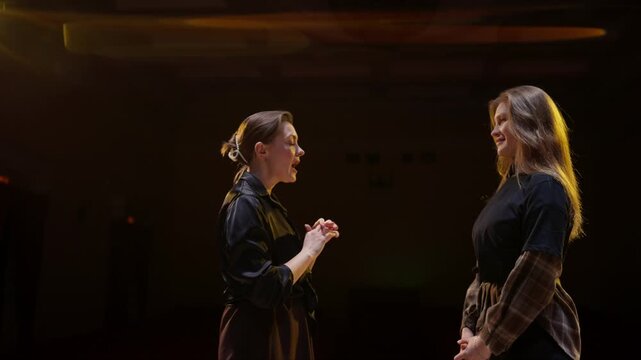 Two women rehearsing expressive dialogue under warm stage lights, practicing performance gestures and emotional delivery in a dramatic theater setting