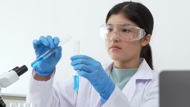 Young Asian women scientist working with test tube in laboratory. Scientist in a lab coat and safety glasses handles a test tube at a desk in a laboratory, make note on laptop, experimentation in a co