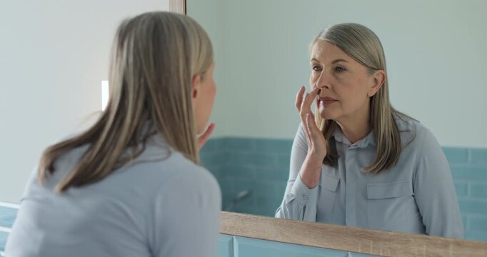 Senior female pleased with her face skin condition in a modern bathroom, anti-aging cosmetics.