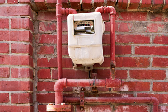 Close-up of an old outdoor gas utility meter encased in white plastic housing, mounted on red brick wall with system of painted metal gas pipe plumbing and valves.