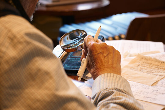 Elderly Expert Meticulously Examining Antique Documents and Digital Records with a Magnifying Glass in an Old Study.