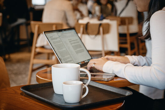 Cozy remote work setting: blurred woman using laptop in bustling cafe, framed by bokeh from foreground coffee mugs.