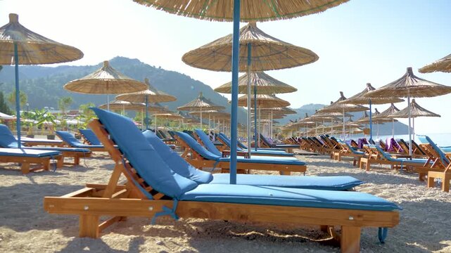 Beach resort with wooden sunbeds and parasols lined along the coast. Bright sunny day with clear water in the background. Travel and tourism concept. Empty sun loungers with blue cushions on a pebble