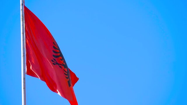 Albanian Flag Waving Proudly Against Blue Sky. National flag of Albania flies high, displaying its black double-headed eagle on a bright red background, under a vibrant sky.
