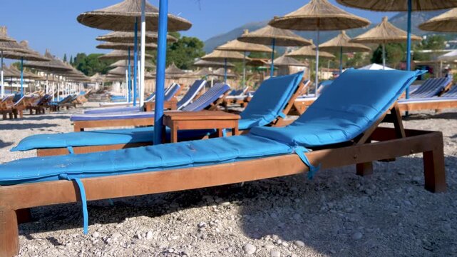 Empty sun loungers with blue cushions on a pebble beach. Rows of straw umbrellas under clear blue sky. Calm seaside resort scene, perfect for summer vacation. Ideal place for relaxation.