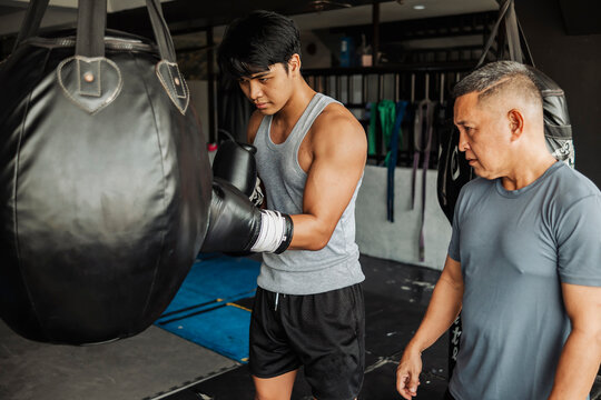 Young Asian man does boxing uppercut drill at the gym, guided by a trainer observing form and technique to improve timing, accuracy, and effective close-range combinations.