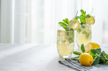 A summer drink with lemon, mint, and ice in a two glasses against a sunny window background. © Kufotos