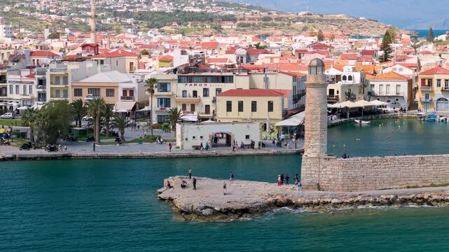 Aerial 70mm view of Rethymno lighthouse at sunny day. Drone flight around Egyptian lighthouse with Venetian Fortezza fortress and old town on background. Scenic Crete harbor landscape, 4k