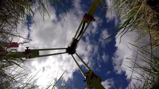 GoPro timelapse from ground looking up at survey tripod with GNSS base station. Clouds moving across blue sky, green grass at frame edges under partly cloudy daylight.