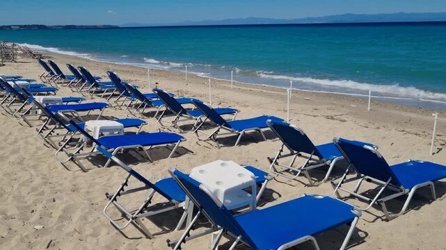 Empty Polychrono Beach on the Kassandra Peninsula in Greece, with blue sun loungers lined along the shore, gentle waves rolling onto the sand under a clear sky in early spring, off season