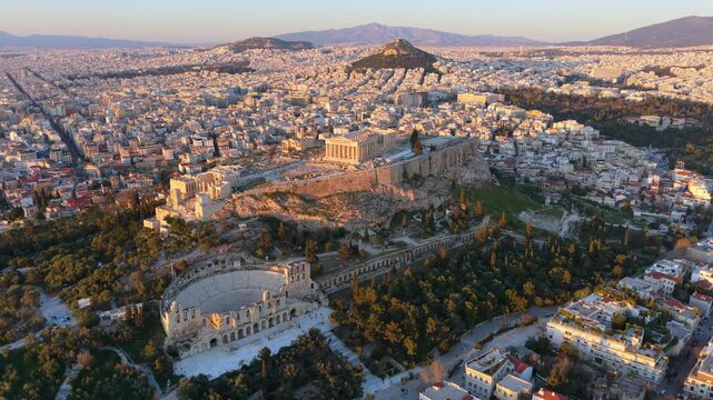 High-angle drone Video of the Parthenon atop the Acropolis hill, overlooking the vast urban sprawl of Athens and Lycabettus Hill at sunset.