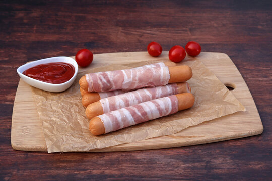 Rohe Berner W&uuml;rstel im Speckmantel mit Ketchup und Tomaten auf Holzbrett.