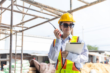 Happy female engineer construction worker stands on site holding tablet using radio for communication wearing safety gear feeling confident with smiling expression during building project © eakgrungenerd