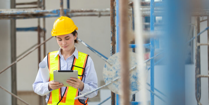 Female construction worker wearing yellow hardhat and safety vest looking at digital tablet with focused expression on building site next to scaffolding and metal framework in daylight outdoors