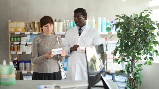Woman choose buy painkiller tablet at pharmacy, compare with other good. Client check for absence of harmful substances, view product in details. African man pharmacist offer available assortment item