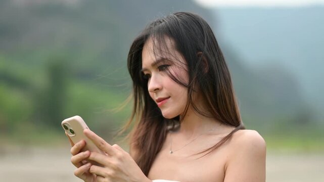 A young woman in a white strapless top is focused on her smartphone outdoors with a blurred natural background.