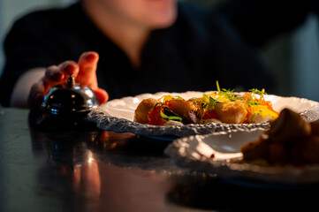 Chef's hand reaching for a silver service bell on a dark counter next to a gourmet plated appetizer in a dimly lit, high-end restaurant kitchen © pavel siamionov