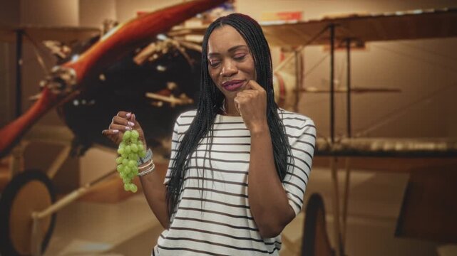Woman holding a bunch of green grapes with hand covering mouth beside a vintage biplane inside museum building; surprise curiosity.