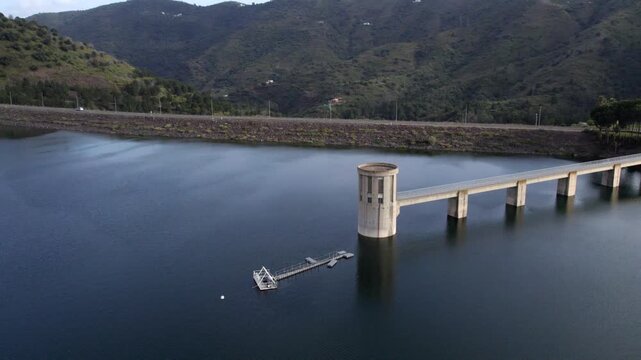Aerial view of a concrete dam intake tower and reservoir with calm water surrounded by green mountains