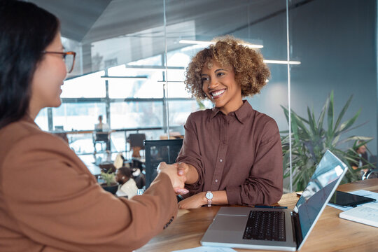 Businesswomen shake hands at desk in modern business office, smiling at meeting. Conveying agreement, woman partnership and professional success. People satisfied with results of team work together
