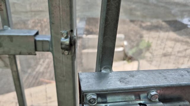 A hand slides a metal barrel bolt latch into place on a galvanized steel gate, securing it against a mesh fence background.