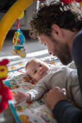 Obraz premium Baby lying on back on play mat while father interacts with colorful toy above, medium close-up framing, bright living room, natural daylight