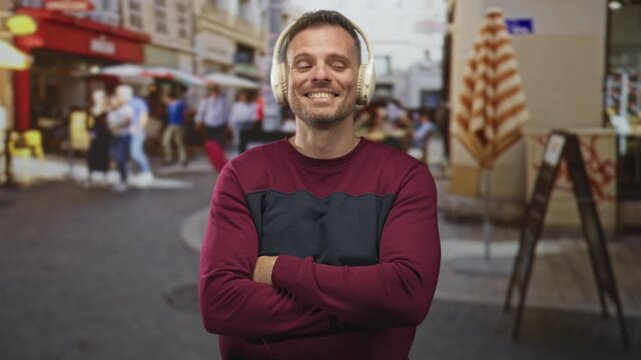 Man with headphones, arms crossed and smiling on a street cafe terrace beside a chalkboard menu and red awning; contentment.