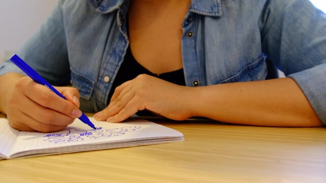 Close up shows hands doodling in a grid notebook with a blue pen on a wooden desk, soft indoor light and shallow focus. Creative scribbles ease stress, helping reset attention after work.