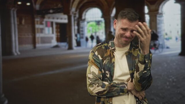 Man soldier in camouflage shirt standing grimacing with hand on forehead inside building archway; frustration fatigue.