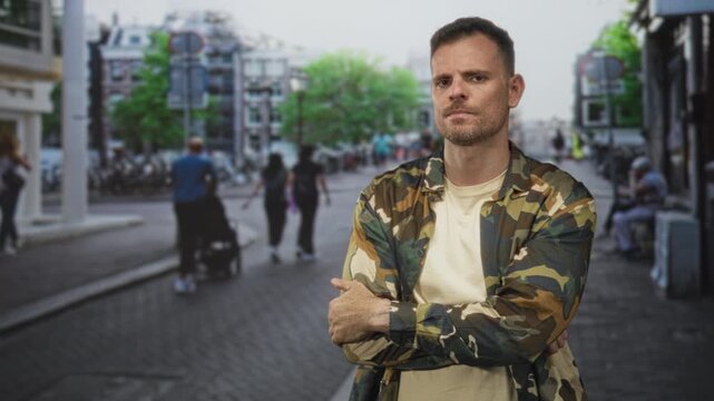 Man soldier in camouflage jacket standing with arms crossed on street beside sidewalk and bicycles, urban scene; pride duty.