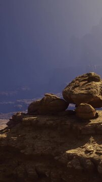 Large, rugged rocks sit gracefully atop a cliff, illuminated by the golden light of dawn. The background shows a vast expanse of mountains and clear blue skies, offering serene beauty.