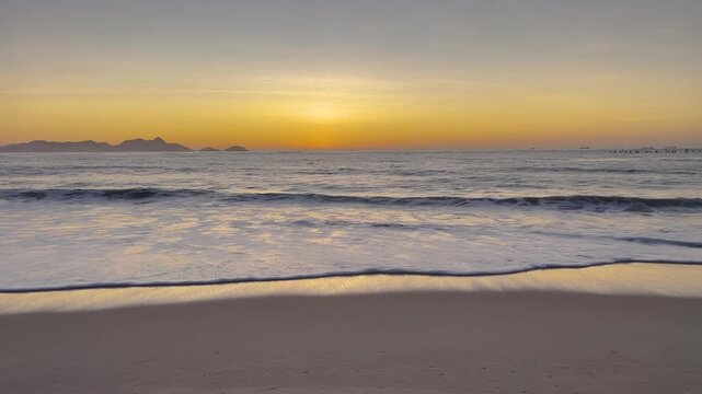 Static shot of golden sunrise over Atlantic Ocean, waves breaking on Copacabana Beach with mountain silhouettes, warm glowing light reflected on wet sand, Rio de Janeiro.
