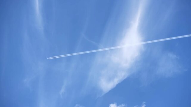 A jet plane creates a contrail against a bright blue sky with wispy clouds. Ideal for travel or aviation-themed projects.
