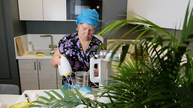 Grandmother with headscarf holding hand mixer and metal flour sifter over blue bowl. Home baking and step by step dough preparation in modern kitchen.