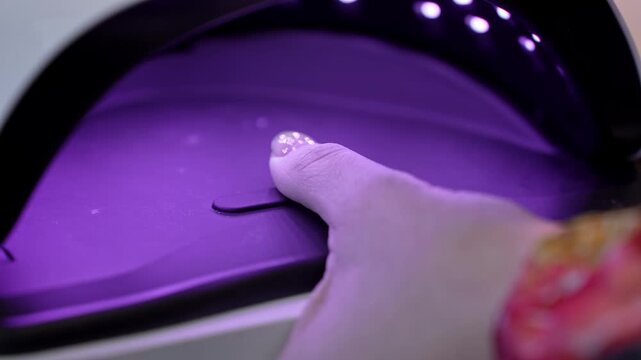 Glitter accent thumb curing under a UV LED nail lamp, glowing in violet light during the final stage of a professional salon manicure.