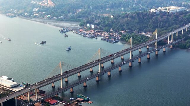 Cinematic aerial view of Atal Setu stretching across the river, with steady traffic, elegant cable pylons, and boats moving through calm waters, framed by lush riverside greenery.