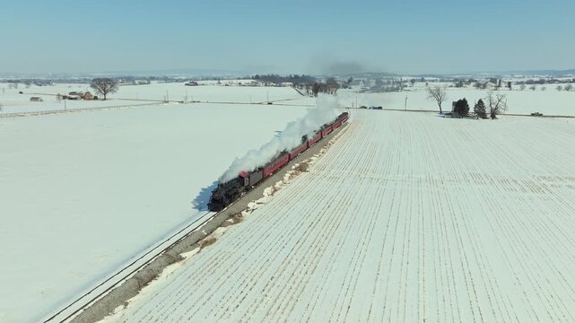 A train moves along tracks through snowy fields. The bright sky shows it's afternoon. The landscape is flat with snow covering the ground. Trees stand in the distance.