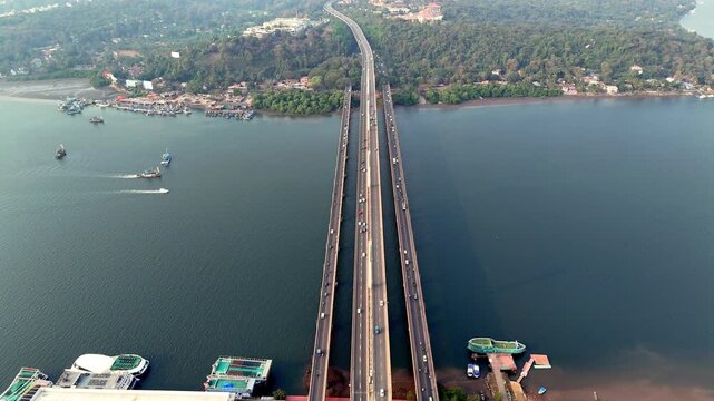 Cinematic 360 degree aerial view of Atal Setu stretching gracefully across a wide river, with elegant cable-stayed towers, flowing traffic, and boats moving below against a calm blue water backdrop.