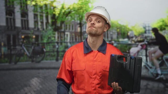 Man builder in hardhat and orange uniform gestures with right hand while holding a black toolbox on street by canal bridge; confidence reliability.