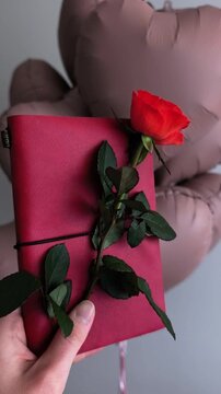 Person's hand holding a beautiful romantic gift. A single red rose is tucked into a red notebook against a background of heart shaped balloons