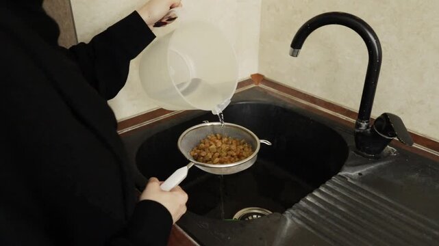 water flows from a plastic pitcher onto soaked raisins held in a mesh strainer over a black sink, showing a practical cleaning stage before adding dried fruit to rice