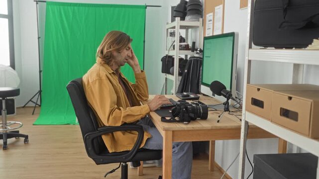 Man photographer types on keyboard with hand to face at computer in studio; concentration creativity.