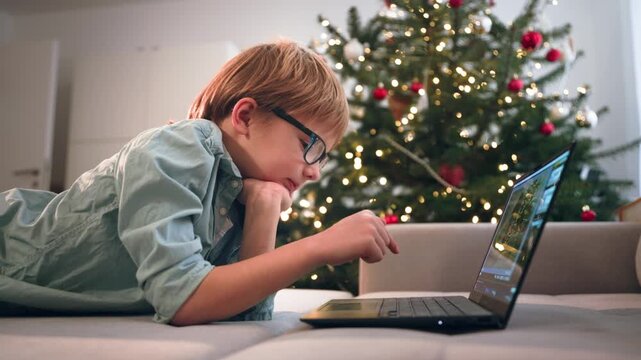 On xmas, a little boy with blond hair and glasses lying on the sofa with laptop