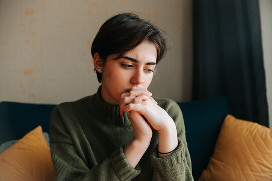 A young adult with short hair sits on a sofa with hands clasped together, appearing deep in thought. The setting includes soft cushions and a cozy atmosphere, suggesting a moment of reflection