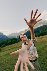 Fototapeta premium A woman in casual outdoor wear extends her hand toward the camera in a mountain meadow, smiling and enjoying a sunny day of active living and exploration in fresh air and wide open sky.