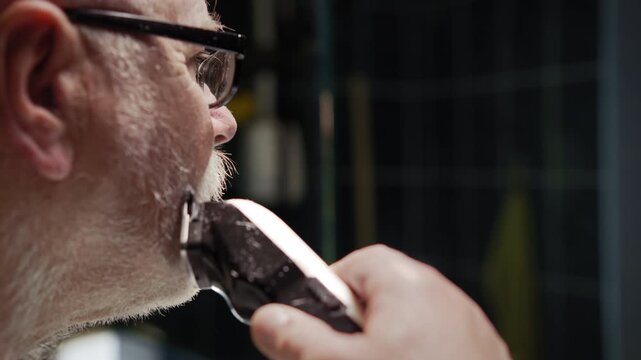 An elderly man shaves his mustache with a trimmer in front of a bathroom mirror