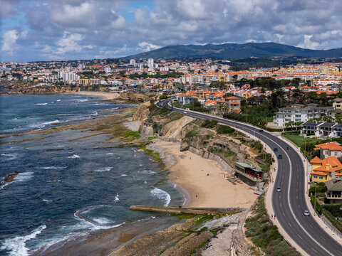 Aerial view of the Avenida Marginal coastal road winding along the rocky shoreline and sandy beach with residential buildings and hills under a cloudy sky in Parede, Lisboa, Portugal.