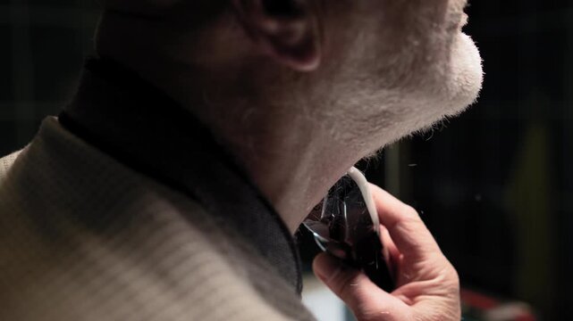 A white haired man shaves with a trimmer in front of a mirror in the bathroom