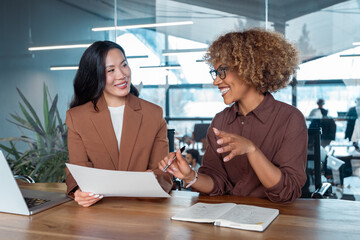 Fototapeta na wymiar Two businesswomen collaborating at modern office desk smiling during meeting with laptop and documents. Multiracial women talking working together. Teamwork and professional communication at workplace